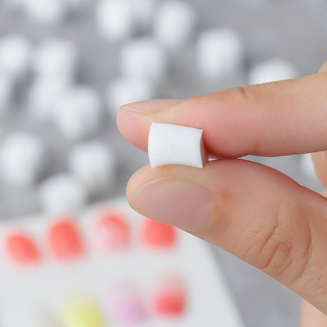 A single sponge cube held between fingers with nail art setup in background.